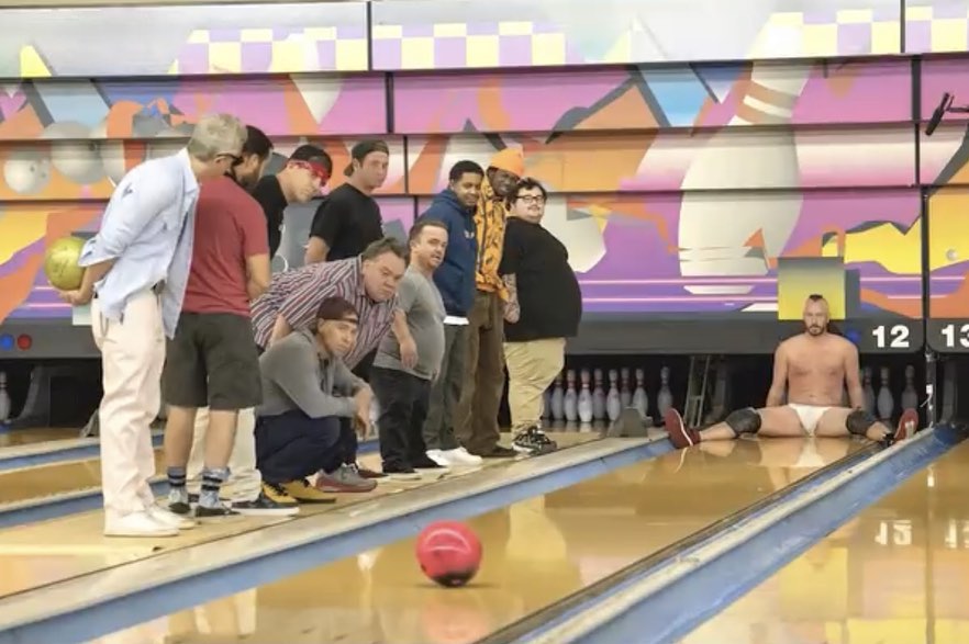 Jackass sitting on the bowling arena with legs wide spread open and bowling ball coming towards him